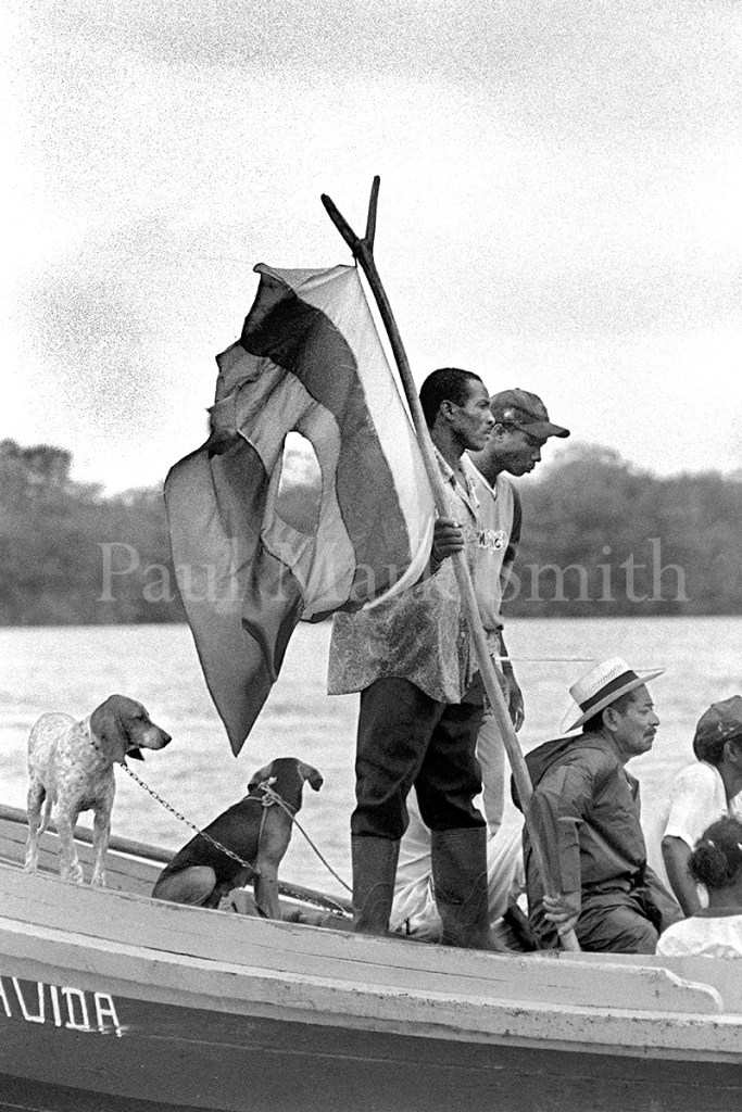 Men and dogs with a flag in a wooden boat that passes along a river