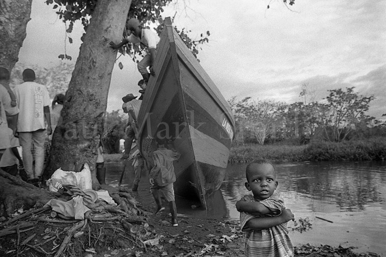 A boy stands on the river's edge by a wooden boat