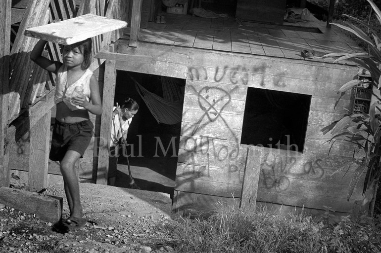 A girl carries a plank on her head and a woman sweeps in a wood house with graffiti threatening death