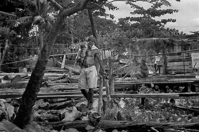 A displaced man carries a small baby along a log