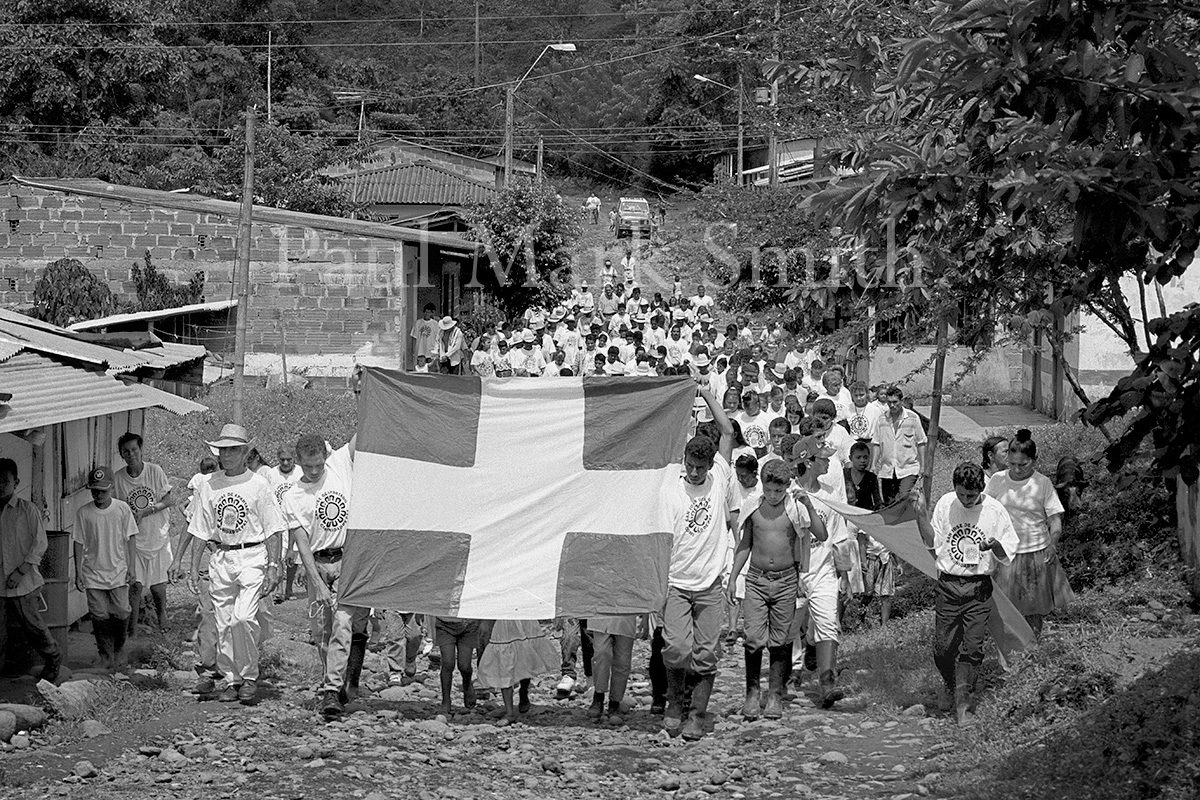 A march of farmers, who hold a flag with a white cross, through a small Colombian village.