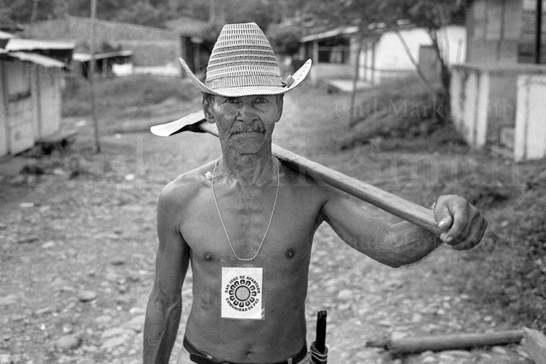 A farmer with an axe and a cardboard tag that reads "San José de Apartadó - Peace Community"