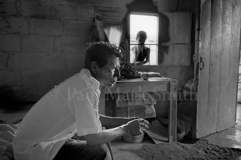 A man sits alone on a bed in a house as someone looks in through the window