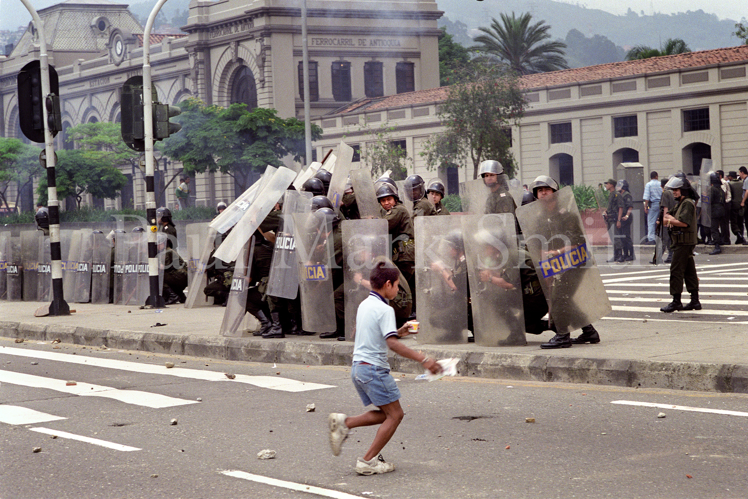 Boy runs across road covered with rocks as police take refuge behind riot shields