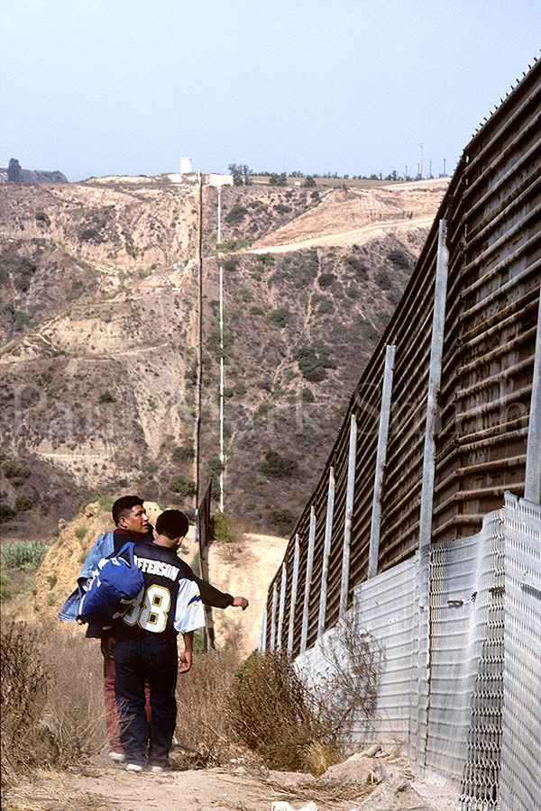 Three migrants walk along the border fence that cuts through dry hills