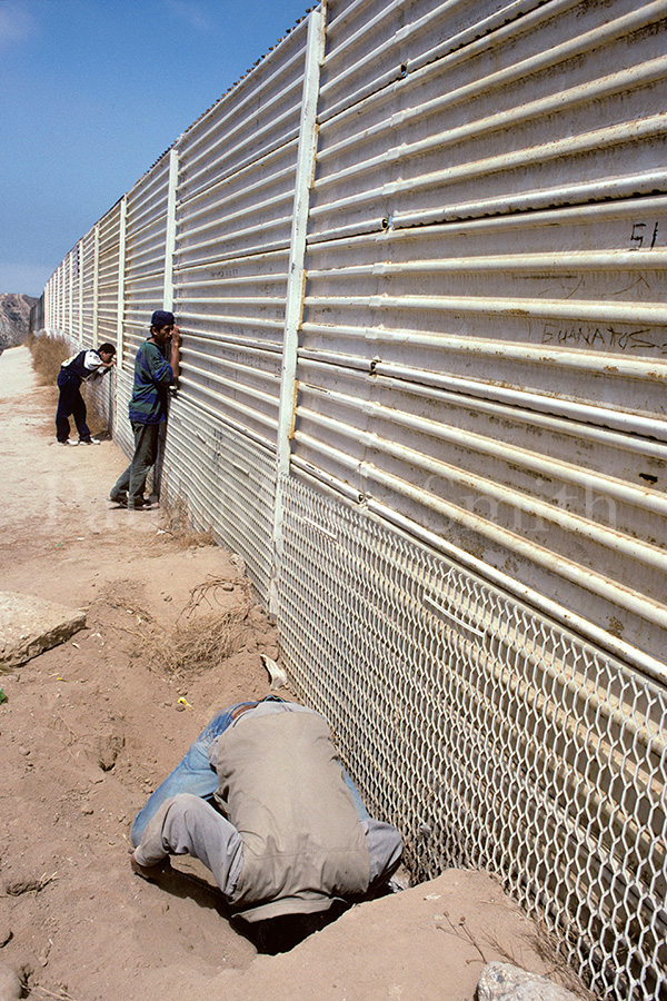 Migrant man sticks his head in the ground to look under a border fence