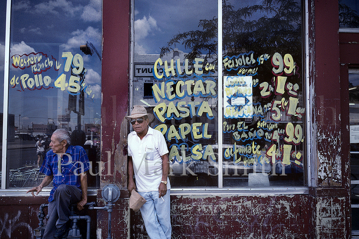 Latino men outside a food store with products and prices written on windows