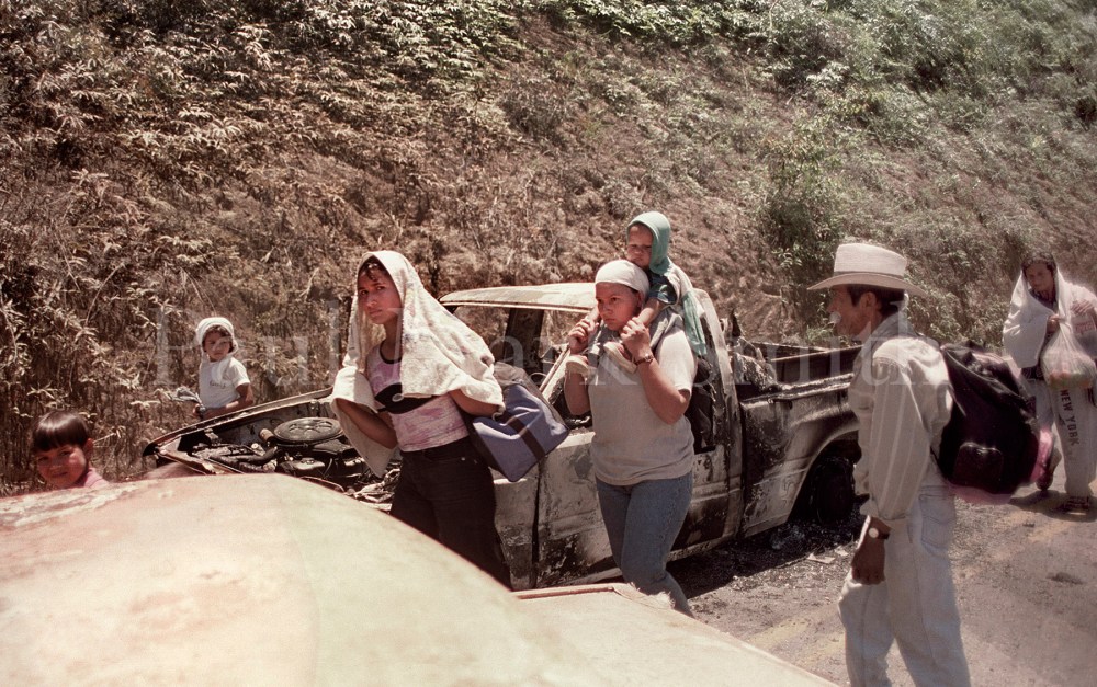 Women, children and an old farmer walk by burnt-out vehicles on a rural road